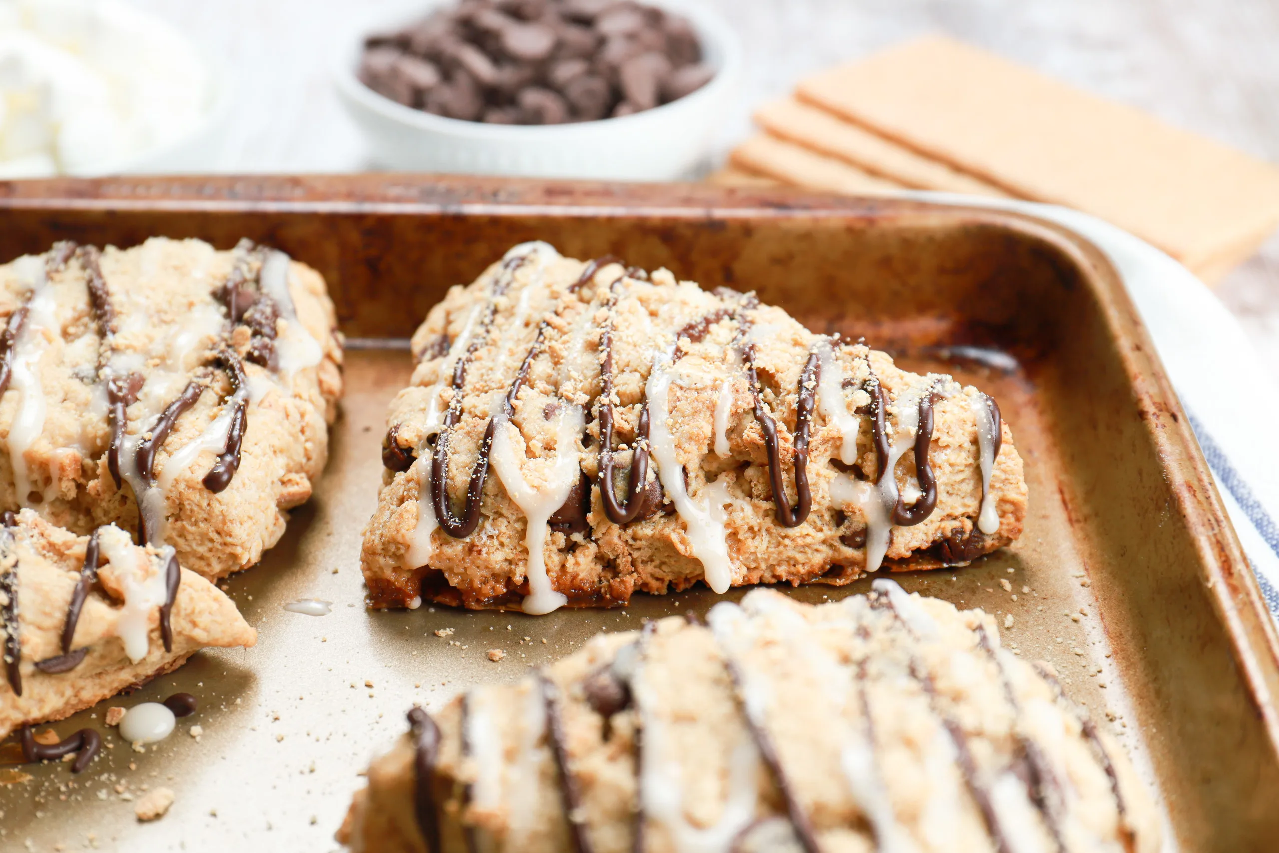 Up close side view of a s'mores scone on a baking sheet with marshmallows, chocolate chips, and graham crackers in the background.